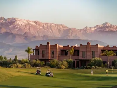 a building with golf carts on a green with mountains in the background at Atlas Gardens Apartment in Sraghna